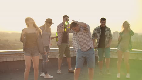 A-company-of-six-young-people-threw-a-party-on-the-roof-with-beer.-Three-young-men-and-two-young-girls-are-dancing-in-plaid-shirts.-Young-blonde-dances-in-the-foreground-in-denim-shorts-and-denim-jacket.
